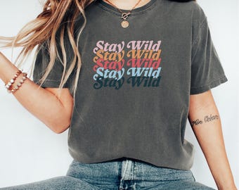 May include: Dark grey t-shirt featuring the text "Stay Wild" in a stacked, retro font. The text is in shades of pink, orange, yellow, red, and blue. The shirt is a casual style, suitable for everyday wear.