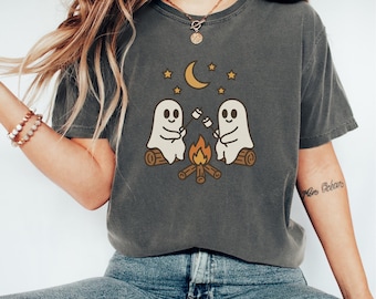 Può includere: T-shirt grigio scuro con un'illustrazione di due fantasmi che arrostiscono marshmallow su un falò. Il design include una luna crescente e stelle, evocando un tema autunnale e accogliente.