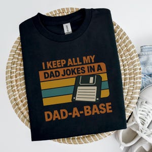 Peut inclure: T-shirt noir avec l'inscription "I KEEP ALL MY DAD JOKES IN A DAD-A-BASE" en orange et blanc. Le design comprend un graphique rétro de disquette. Le t-shirt est présenté sur un panier en osier.