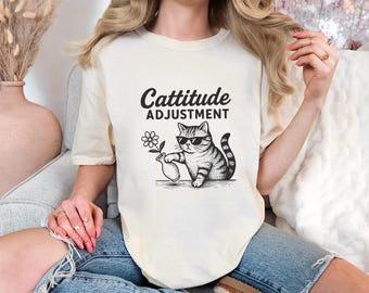 May include: Cream-colored t-shirt with the text "Cattitude Adjustment" above a graphic of a cat wearing sunglasses and holding a vase with a flower. The cat is black and white striped.