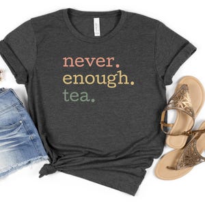 以下が含まれることがあります： ピンク、イエロー、グリーンのグラデーションでレトロなフォントで「never. enough. tea.」とプリントされたダークヘザーグレーのTシャツです。