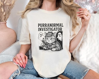 May include: Cream-colored t-shirt featuring a black and white graphic of a cat detective with a magnifying glass and two ghosts. The text "PURRANORMAL INVESTIGATOR" is printed above the graphic.