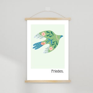 Può includere: Una stampa artistica incorniciata con un uccello verde stilizzato con motivi floreali e a foglie, piume della coda blu e fiori rosa. La parola "Frieden" è stampata in nero in basso. La stampa è tenuta da una cornice magnetica in legno.