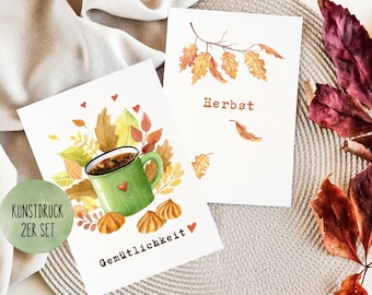 May include: Two watercolor illustrations on white cardstock. The first illustration features a green mug with coffee, surrounded by fall leaves and the text "Gemütlichkeit" and "KUNSTRUCK 2ER SET". The second illustration features a branch with fall leaves and the text "Herbst".