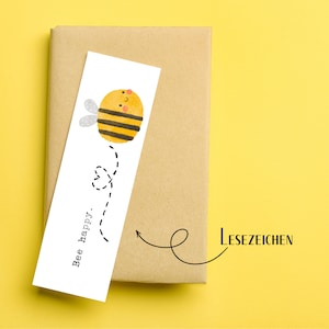 Peut inclure: Un marque-page blanc avec une illustration d'abeille jaune et noire et le texte "Bee happy." Le marque-page est attaché à un paquet en papier brun avec le mot "LESEZEICHEN" écrit dessus.
