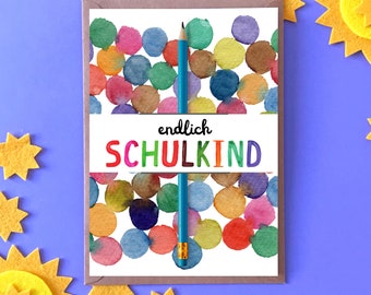 May include: A card with a colourful watercolour background of circles and the text "endlich SCHULKIND" in rainbow colours. A blue pencil is leaning against the card.