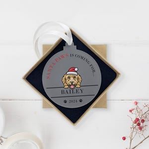May include: A gray round Christmas ornament with a dog wearing a Santa hat. The ornament says "Santa Paws is coming for..." and "Bailey 2021".