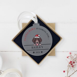 May include: A gray round Christmas ornament with a dog wearing a Santa hat. The ornament says "Santa Paws is coming for... Bailey 2021".