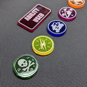 May include: A collection of colorful game tokens and markers. Includes a rectangular token with "ABILITY USED" text, and round tokens with various symbols like a skull and crossbones, lightning bolt, and stylized letters.