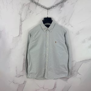 Könnte beinhalten: Langärmliges Hemd mit kleinem weiß-blauem Karomuster. Das Hemd hat einen Button-Down-Kragen und ein kleines gesticktes Logo auf der Brust. Es hängt an einem schwarzen Kleiderbügel.