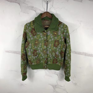 Puede incluir: Chaqueta verde con estampado floral, cuello de forro polar verde y puños de canalé. La chaqueta tiene cierre de botones en la parte delantera y dos bolsillos delanteros. El estampado floral incluye detalles en marrón, amarillo y gris. La chaqueta está colgada en una percha de madera.