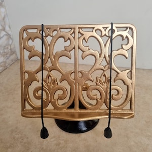 May include: A gold-coloured metal book stand featuring an intricate scrollwork design. The stand includes a horizontal ledge for resting books and two thin black elastic cords with weighted ends hanging down from the top to hold pages open.