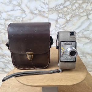 May include: A vintage Specto 88 Colorshot movie camera with a grey textured body, a silver lens, and a brown leather carrying case. The camera features a wrist strap and a front panel with yellow circular indicators and black text labels.