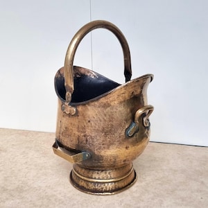 May include: A vintage brass coal scuttle. It features a curved handle and a hammered texture. The scuttle has a rounded base and a flared top, with small handles on the sides. The interior is dark, contrasting with the polished brass exterior.