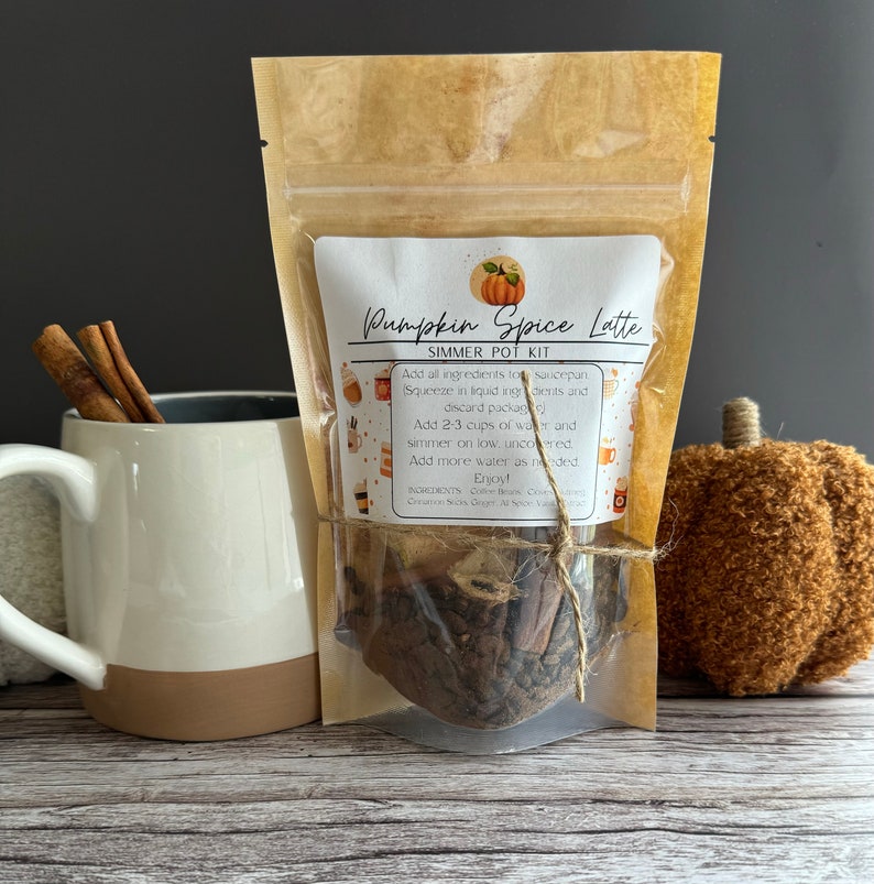 May include: A brown paper pouch with a label that reads "Pumpkin Spice Latte Simmer Pot Kit". The pouch contains a mix of spices and coffee beans. The pouch is tied with twine and there is a white mug and a brown plush pumpkin in the background.