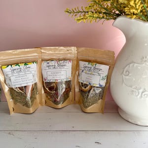 May include: Three small, brown paper pouches labeled "Spring Clean," "Spring Blossom," and "Spring Breeze." Each pouch contains dried herbs and botanicals. A white vase with yellow flowers is in the background.
