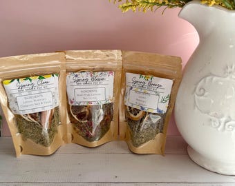 May include: Three small, brown paper pouches labeled "Spring Clean," "Spring Blossom," and "Spring Breeze." Each pouch contains dried herbs and botanicals. A white vase with yellow flowers is in the background.