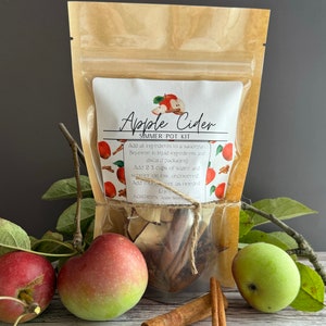May include: A brown paper bag with a clear plastic window containing a simmer pot kit for apple cider. The kit includes apple slices, cinnamon sticks, and other spices. The bag is tied with twine and has a label that reads "Apple Cider Simmer Pot Kit".