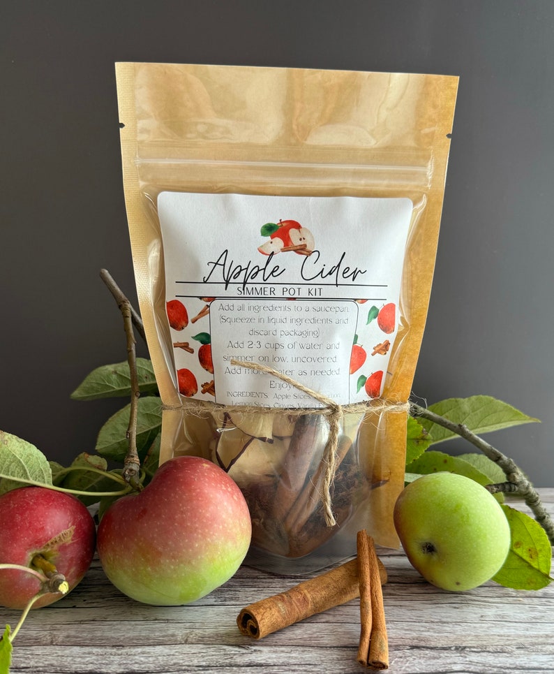 May include: A brown paper bag with a clear plastic window containing a simmer pot kit for apple cider. The kit includes apple slices, cinnamon sticks, and other spices. The bag is tied with twine and has a label that reads "Apple Cider Simmer Pot Kit".