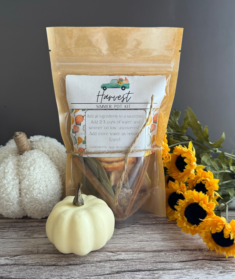 May include: A brown paper bag with a label that says "Harvest Simmer Pot Kit". The bag contains dried apple slices, cinnamon sticks, and other spices. The bag is tied with twine and decorated with a small truck illustration.