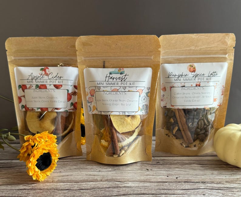May include: Three brown paper bags with clear plastic windows containing mini simmer pot kits. The bags are labeled "Apple Cider", "Harvest", and "Pumpkin Spice Latte". Each bag contains a variety of ingredients, including dried fruit, spices, and herbs.