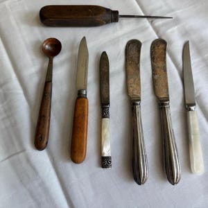May include: Collection of antique cutlery and tools, including a wooden-handled awl, a small wooden spoon, and several knives. The knives feature wooden, silver, and mother-of-pearl handles. The items are arranged on a white cloth.