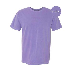 Pode incluir: Uma t-shirt de manga curta em cor violeta s&oacute;lida com decote redondo. Um gr&aacute;fico circular roxo no canto superior direito cont&eacute;m o texto branco Violet. A pe&ccedil;a apresenta um corte padr&atilde;o com uma bainha reta.