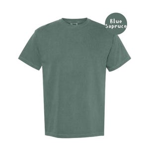 Pode incluir: Uma t-shirt de manga curta com gola redonda num tom azul abeto s&oacute;lido e suave. A pe&ccedil;a apresenta um corte padr&atilde;o com gola canelada e uma textura de tecido lisa e consistente. Um emblema circular no canto superior direito exibe o texto Blue Sapruce.