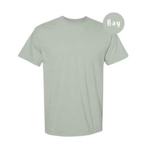 Pode incluir: Uma t-shirt de manga curta com gola redonda num tom s&oacute;lido de verde s&aacute;lvia claro. A pe&ccedil;a apresenta um corte padr&atilde;o com gola canelada e mangas com bainha. Um emblema circular no canto superior direito exibe o texto Bay em letras brancas.