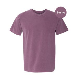 Pode incluir: Uma t-shirt de manga curta com gola redonda numa cor roxa baga s&oacute;lida. O tecido tem uma textura ligeiramente lavada e mate. Um emblema circular roxo no canto superior direito cont&eacute;m o texto branco Berry, indicando a cor da pe&ccedil;a.