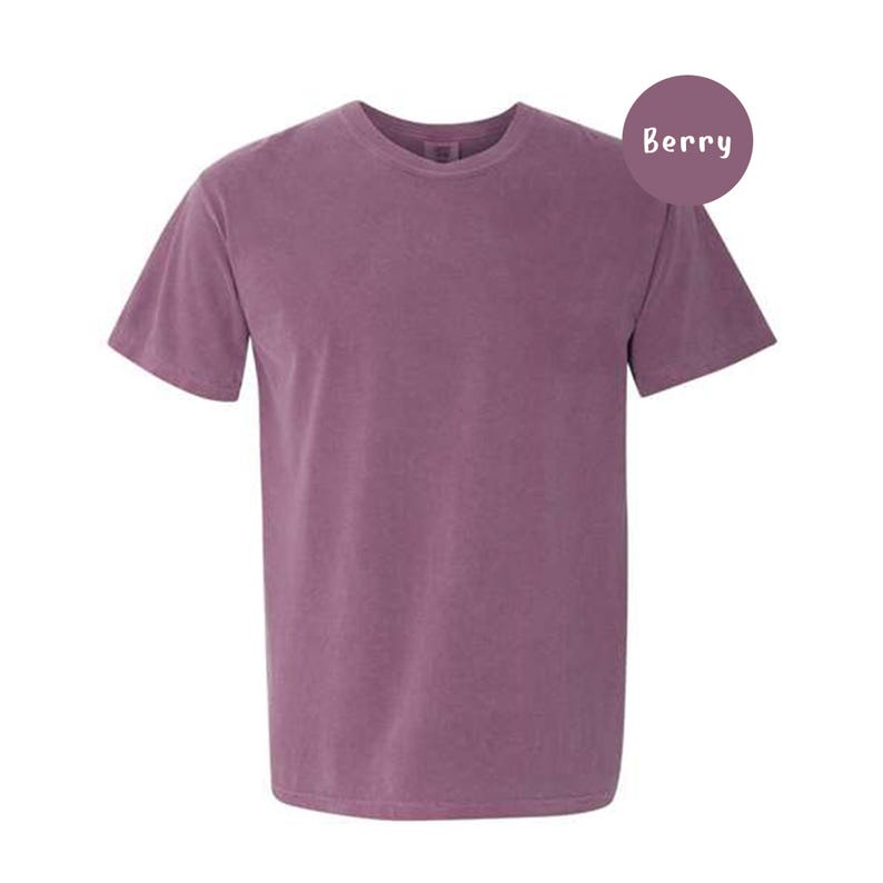 Pode incluir: Uma t-shirt de manga curta com gola redonda numa cor roxa baga s&oacute;lida. O tecido tem uma textura ligeiramente lavada e mate. Um emblema circular roxo no canto superior direito cont&eacute;m o texto branco Berry, indicando a cor da pe&ccedil;a.