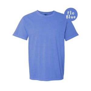 Pode incluir: Uma t-shirt de manga curta com gola redonda numa cor azul pervinca s&oacute;lida. O tecido tem um aspeto ligeiramente lavado e mate. Um gr&aacute;fico circular azul no canto superior direito cont&eacute;m o texto branco Flo Blue, indicando a cor.