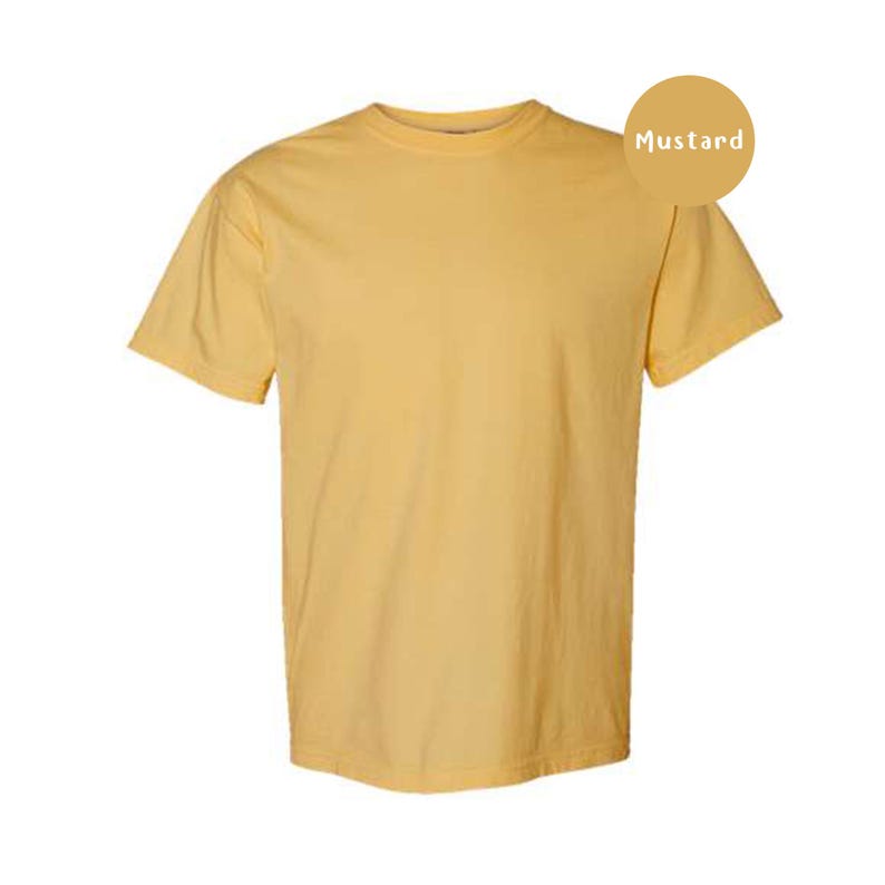 Pode incluir: Uma t-shirt de manga curta em amarelo mostarda s&oacute;lido com decote redondo. A pe&ccedil;a apresenta um design simples e sem padr&otilde;es num tom amarelo claro. Uma etiqueta circular no canto superior direito exibe o texto Mustard em letras brancas.