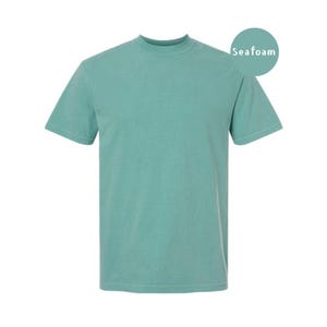 Pode incluir: Uma t-shirt de manga curta com gola redonda numa cor verde espuma do mar s&oacute;lida. A pe&ccedil;a apresenta um corte padr&atilde;o com gola canelada e mangas com bainha. Um emblema circular no canto superior direito exibe o texto Seafoam em branco.