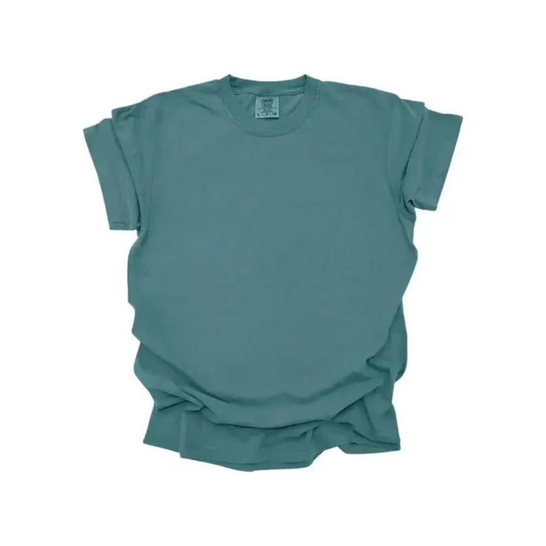 K&ouml;nnte beinhalten: Ein einfarbiges, teal-farbenes T-Shirt mit kurzen &Auml;rmeln. Das Shirt besteht aus einem weichen Material und hat einen Rundhalsausschnitt. Die &Auml;rmel sind hochgekrempelt. Das Shirt liegt flach auf einer wei&szlig;en Oberfl&auml;che.