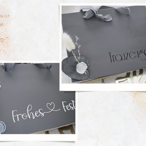May include: Two gray gift bags with white lettering. One bag says "Frohes Fest" with a heart symbol. The other bag says "Trauzeuge".