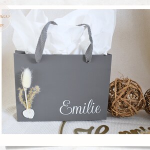 May include: A gray gift bag with white tissue paper inside. The bag has a white handle and the name "Emilie" is printed on the front in white lettering. There is a small dried flower arrangement on the front of the bag.