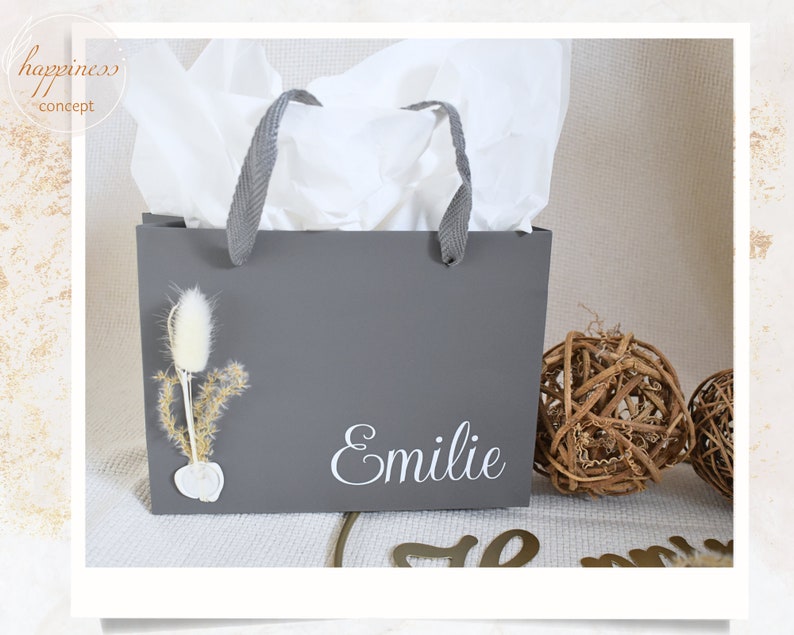May include: A gray gift bag with white tissue paper inside. The bag has a white handle and the name "Emilie" is printed on the front in white lettering. There is a small dried flower arrangement on the front of the bag.