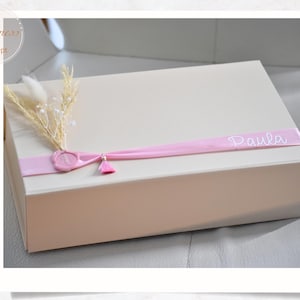 Può includere: Una confezione regalo rettangolare color crema, adornata con un nastro rosa con il nome "Paula". La scatola è impreziosita da fiori secchi, un sigillo di cera rosa e un piccolo nappa, suggerendo un regalo personalizzato.