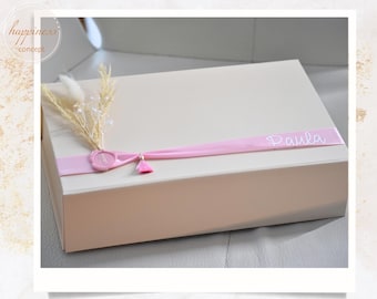 May include: A rectangular gift box in a neutral tone, adorned with a pink ribbon bearing the name "Paula." The box is embellished with a decorative arrangement of dried flowers, a pink wax seal, and a small tassel, suggesting a personalized gift.