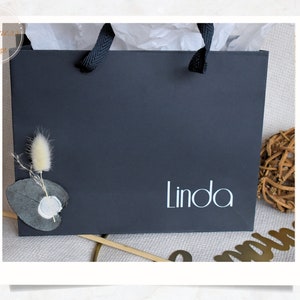 Può includere: Una borsa regalo nera con un manico di carta bianca e la parola "Linda" stampata in bianco sul davanti. La borsa è decorata con un piccolo rametto di fiori secchi e una foglia di eucalipto.