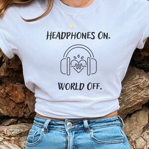 May include: White t-shirt with the text "Headphones On. World Off." in black. The design includes a graphic of headphones with a heart and sound wave in the center. The shirt is paired with blue jeans.
