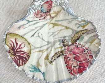 May include: A decorative seashell with a silver-painted edge. The interior features a watercolor design of a sea turtle, nautilus, and coral in shades of pink, green, and blue. The background has a white base with green and blue seaweed.