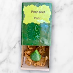 Op de afbeelding: Een groen, peervormig object ligt in een kleine doos gevuld met bruin papier. De doos is gedeeltelijk open en onthult de peer. De doos heeft een groen deksel met de tekst "Pear-fect Pear".