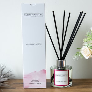 May include: A white box with black text that reads "Clyde Candles Scotland" and "Cranberry & Apple". The box is next to a clear glass bottle with a silver lid and black reeds. The bottle has a label that reads "Clyde Candles Scotland" and "Cranberry & Apple".