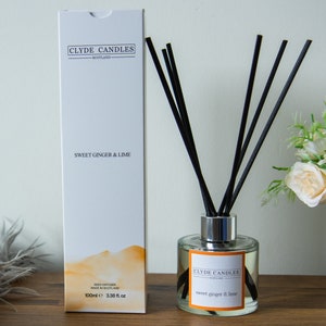 May include: A white box with black text that reads "CLYDE CANDLES SCOTLAND" and "SWEET GINGER & LIME". The box is next to a clear glass bottle with a silver lid and black reeds. The bottle is filled with a light yellow liquid and has a label that reads "CLYDE CANDLES SCOTLAND" and "sweet ginger & lime".