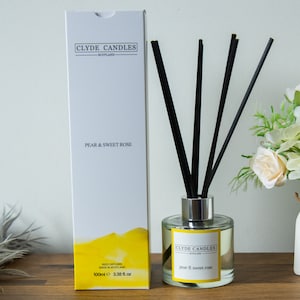 May include: A white box with a yellow and white design and the text "Clyde Candles Scotland" and "Pear & Sweet Rose". A clear glass bottle with a silver lid and black reeds is filled with a yellow liquid. The bottle has a label that reads "Clyde Candles Scotland" and "Pear & Sweet Rose".