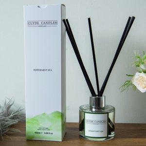 May include: A white box with black text that reads "Clyde Candles - Scotland" and "Peppermint Spa". The box is next to a clear glass bottle with a silver lid and black reeds. The bottle has a label that reads "Clyde Candles - Scotland" and "Peppermint Spa".
