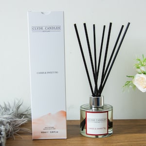 May include: A white box with the text "Clyde Candles Scotland" and "Cassis & Sweet Fig" on it. The box is next to a clear glass bottle with black reeds and the text "Clyde Candles Scotland" and "Cassis & sweet fig" on the label. The bottle is on a wooden surface.