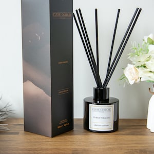 May include: A black and gold diffuser bottle with black reeds and a label that reads "Clyde Candles" and "Cuban Tobacco Limited Edition". The bottle is sitting on a wooden surface.